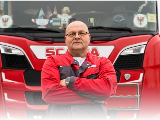 Ein ZEKU-Fahrer in roter Arbeitsjacke steht mit verschränkten Armen vor der Front seiner roten Scania-Zugmaschine.